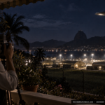 Illustration of a man photographing a motionless, light-reflecting object over a military installation from a Rio de Janeiro veranda at night, December 9, 1954
